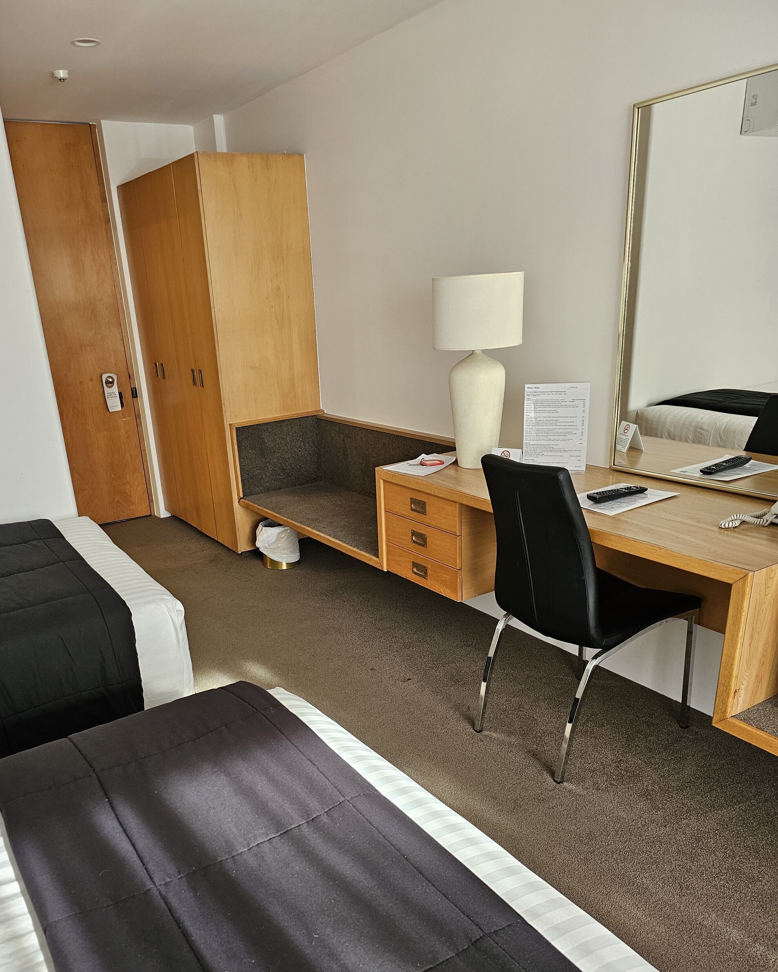 A hotel room with a bed, a desk with a chair, a wardrobe, a mirror, and a lamp.