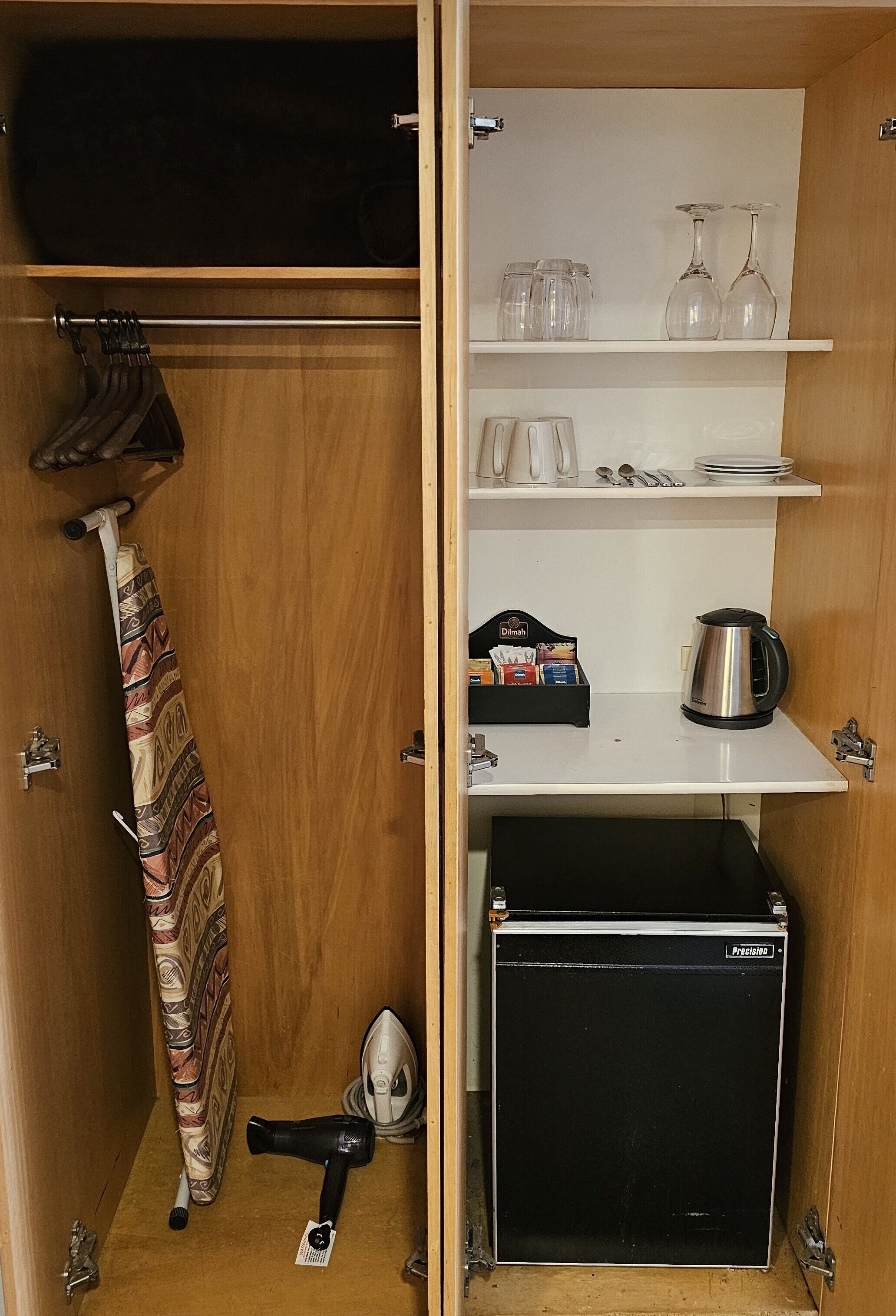 A compact storage unit with shelves holding glassware, a kettle, and a vacuum cleaner.