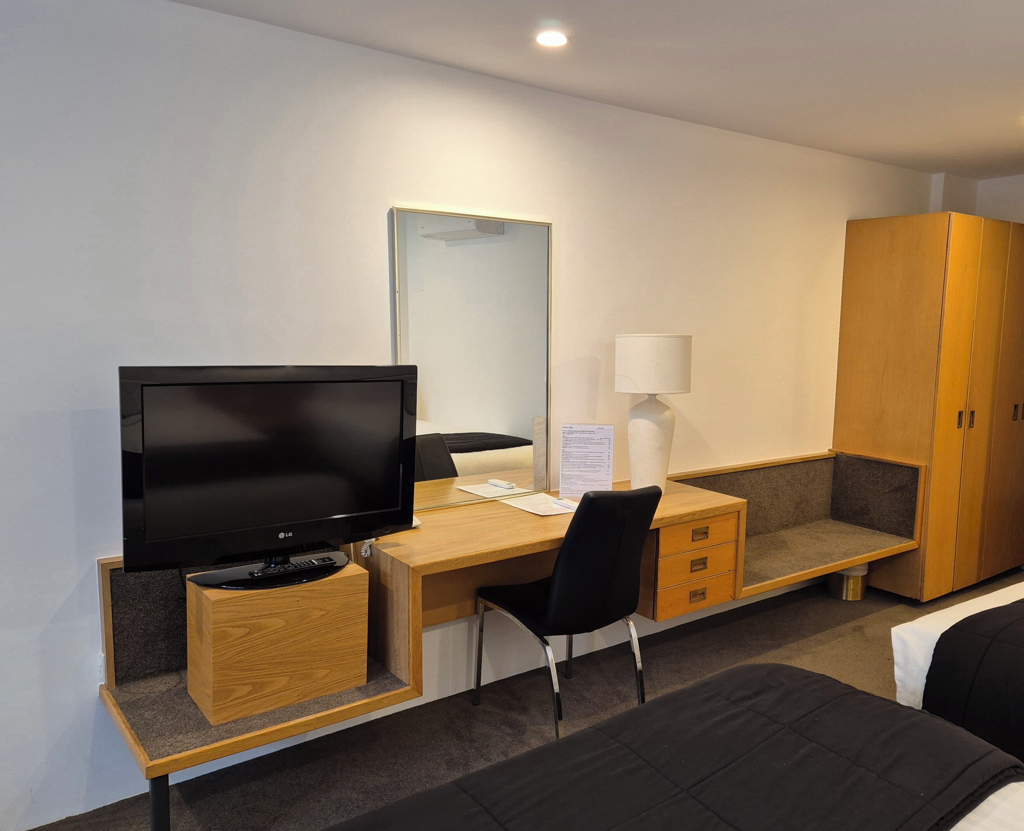 A hotel room with a wooden desk, a television, a chair, a bed, a wardrobe, and a bedside lamp.