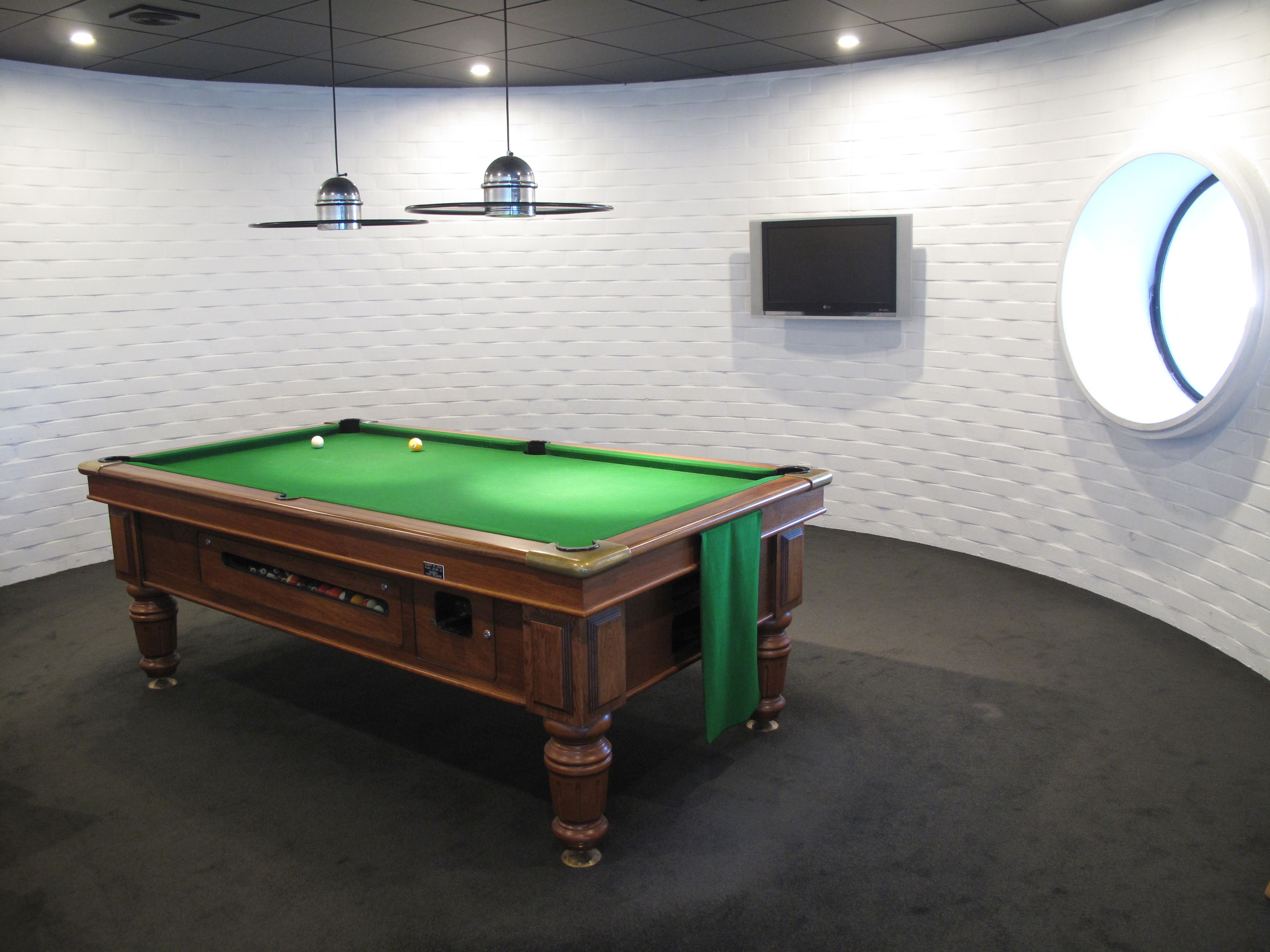 A pool table in a room with white brick walls, a flat-screen TV, and a round mirror.