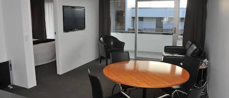 A conference room with a round wooden table, black chairs, a flat-screen TV, and a balcony with a view.