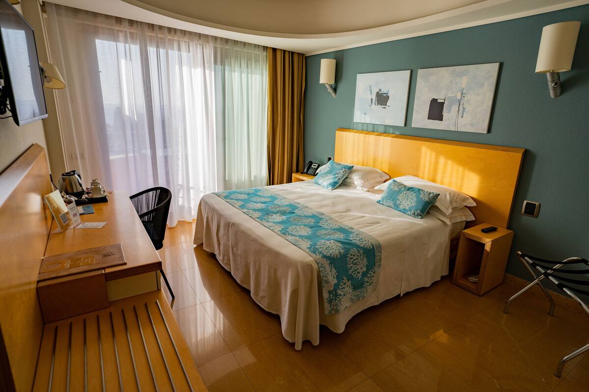 A hotel room with a bed, desk, chair, and a small table with a lamp.