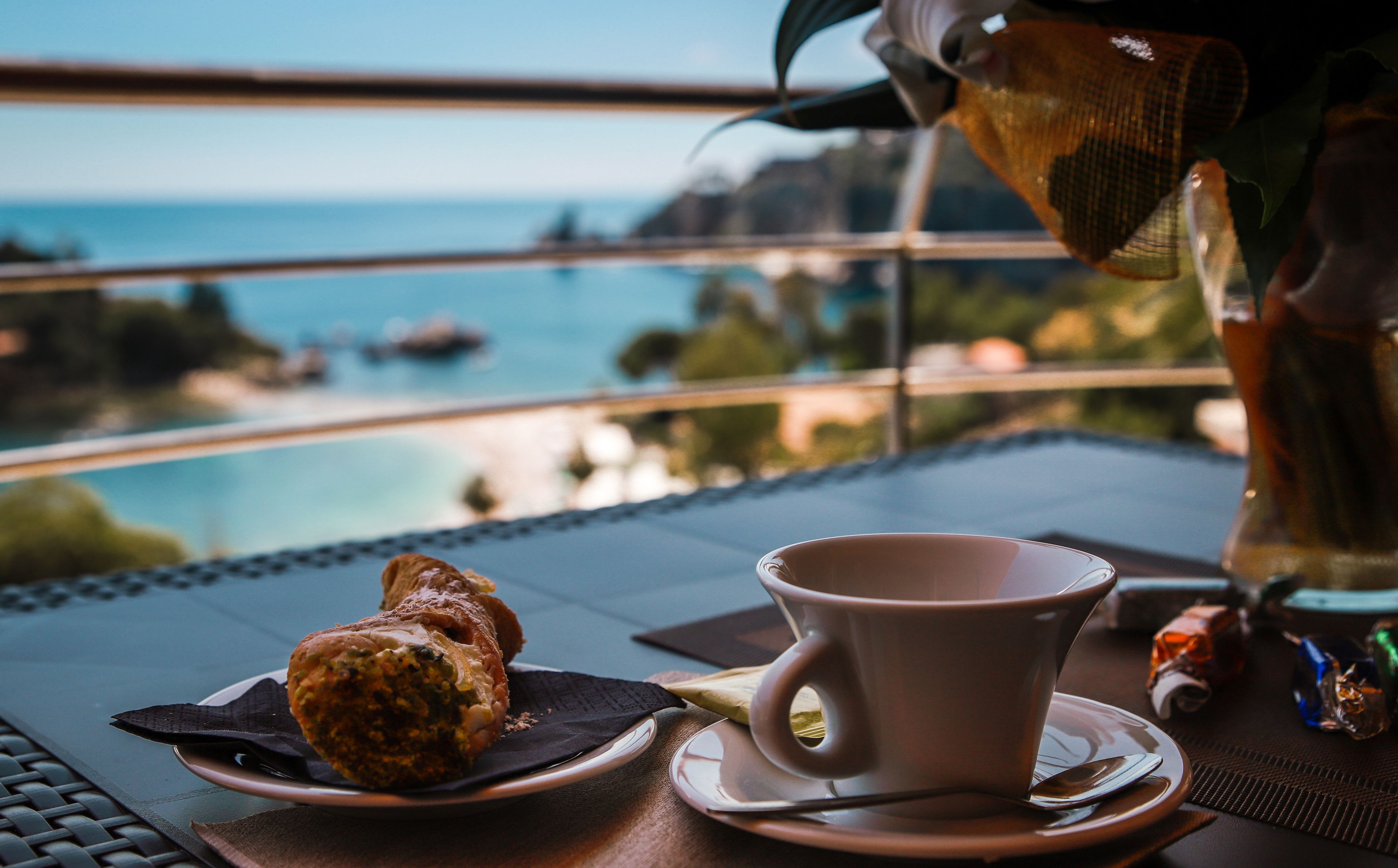 A table with a cup and a pastry, overlooking a scenic view of the sea and boats.