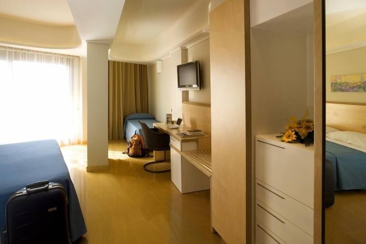 A hotel room with a bed, a desk with a computer, a television, and a suitcase.