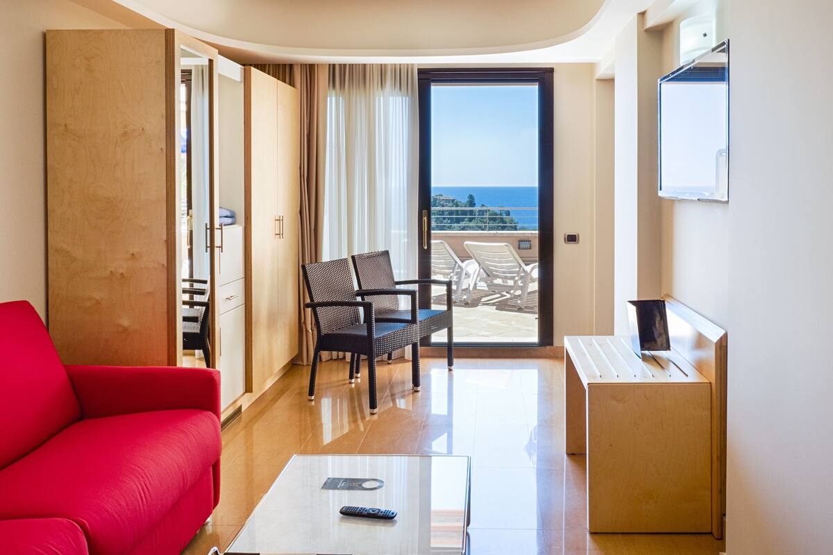 A hotel room with a red sofa, a small table with a remote control, a chair, and a balcony with a view of the sea.