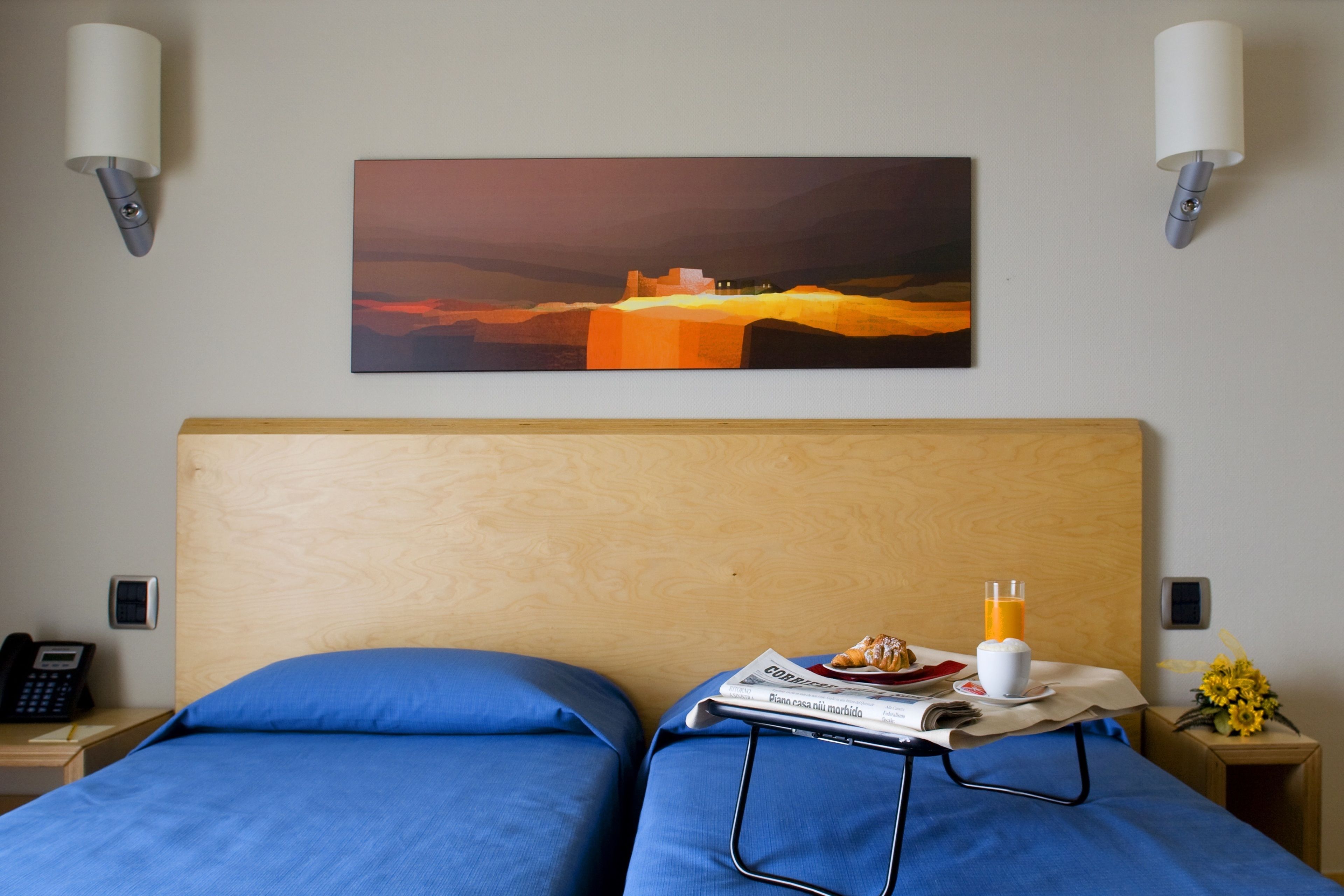 A hotel room with two beds, a painting on the wall, a bedside table with a phone, a glass of orange juice, and a newspaper.