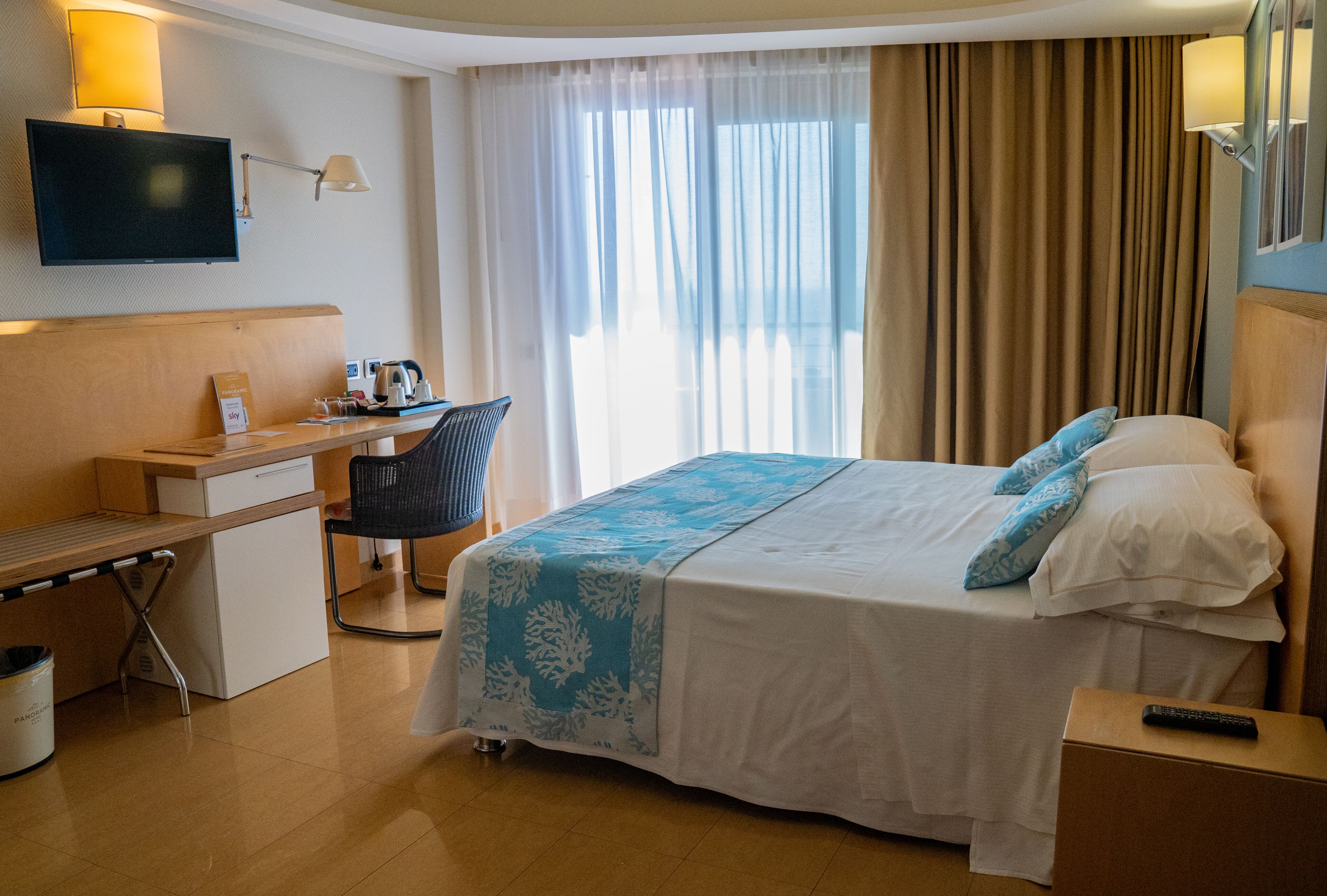Una habitación de hotel con cama, escritorio, silla, televisor y ventana con cortinas.