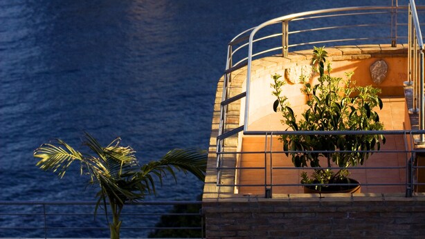 Vista costera con un edificio que domina el mar, un balcón con planta en maceta y una isla rocosa al fondo.
