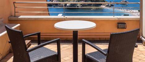 A balcony with wicker chairs and a small table, overlooking a coastal area with rocky outcrops and boats.