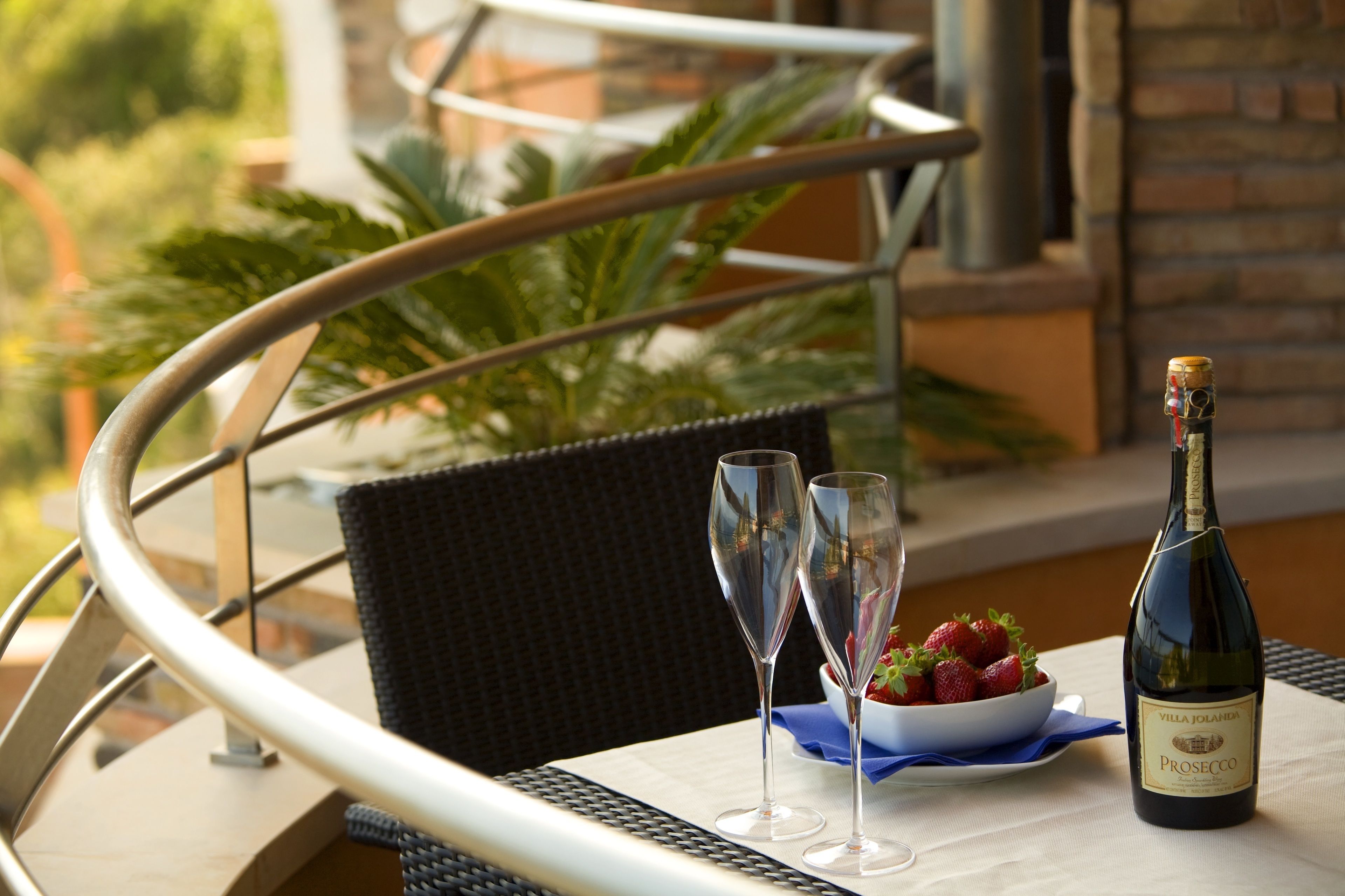 Una mesa con una botella de Prosecco, dos copas de vino y un cuenco de fresas en una terraza con una silla de mimbre y barandilla de metal.