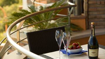 Una mesa con una botella de Prosecco, dos copas de vino y un cuenco de fresas en una terraza con una silla de mimbre y barandilla de metal.