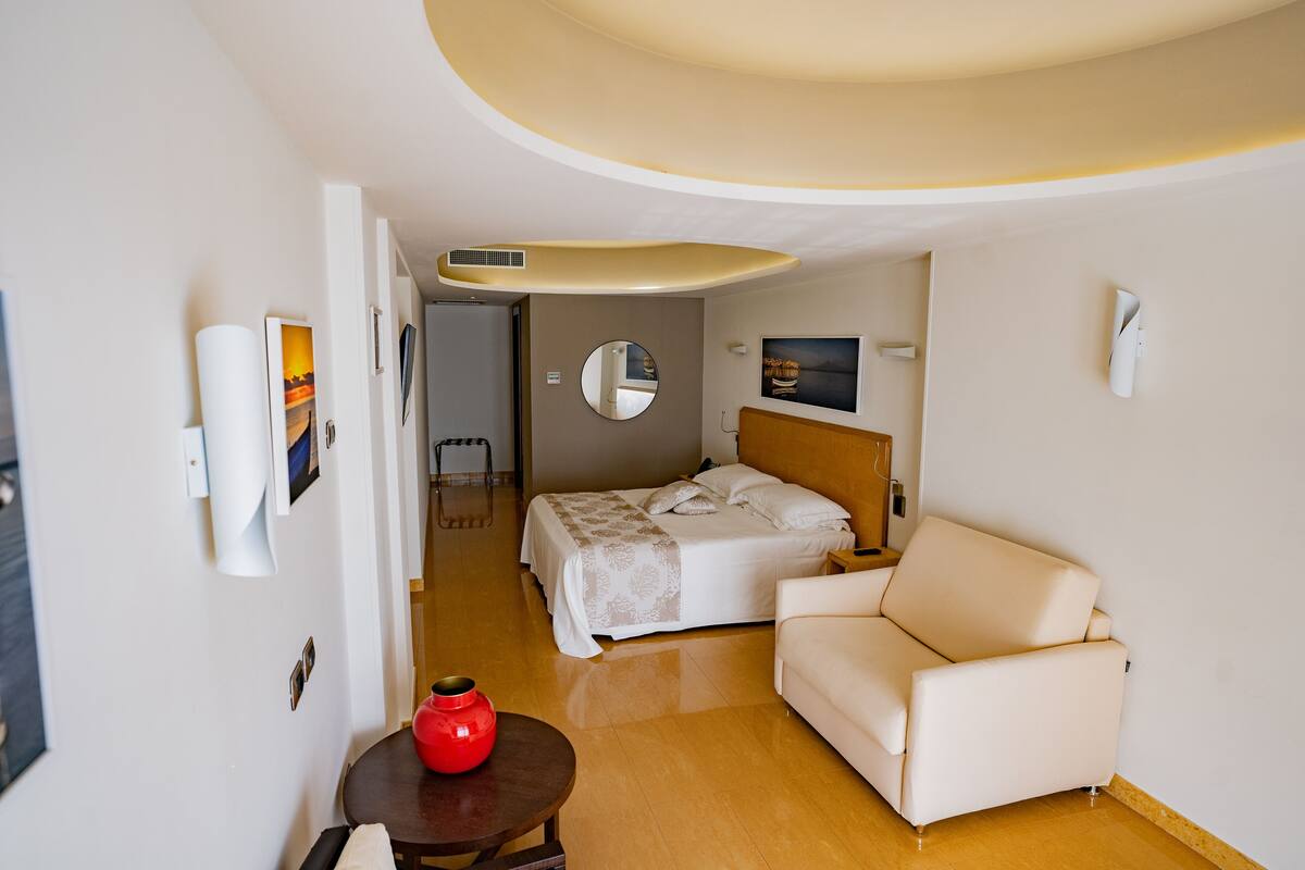 A hotel room with a bed, a sofa, a small table with a red vase, and a mirror on the wall.