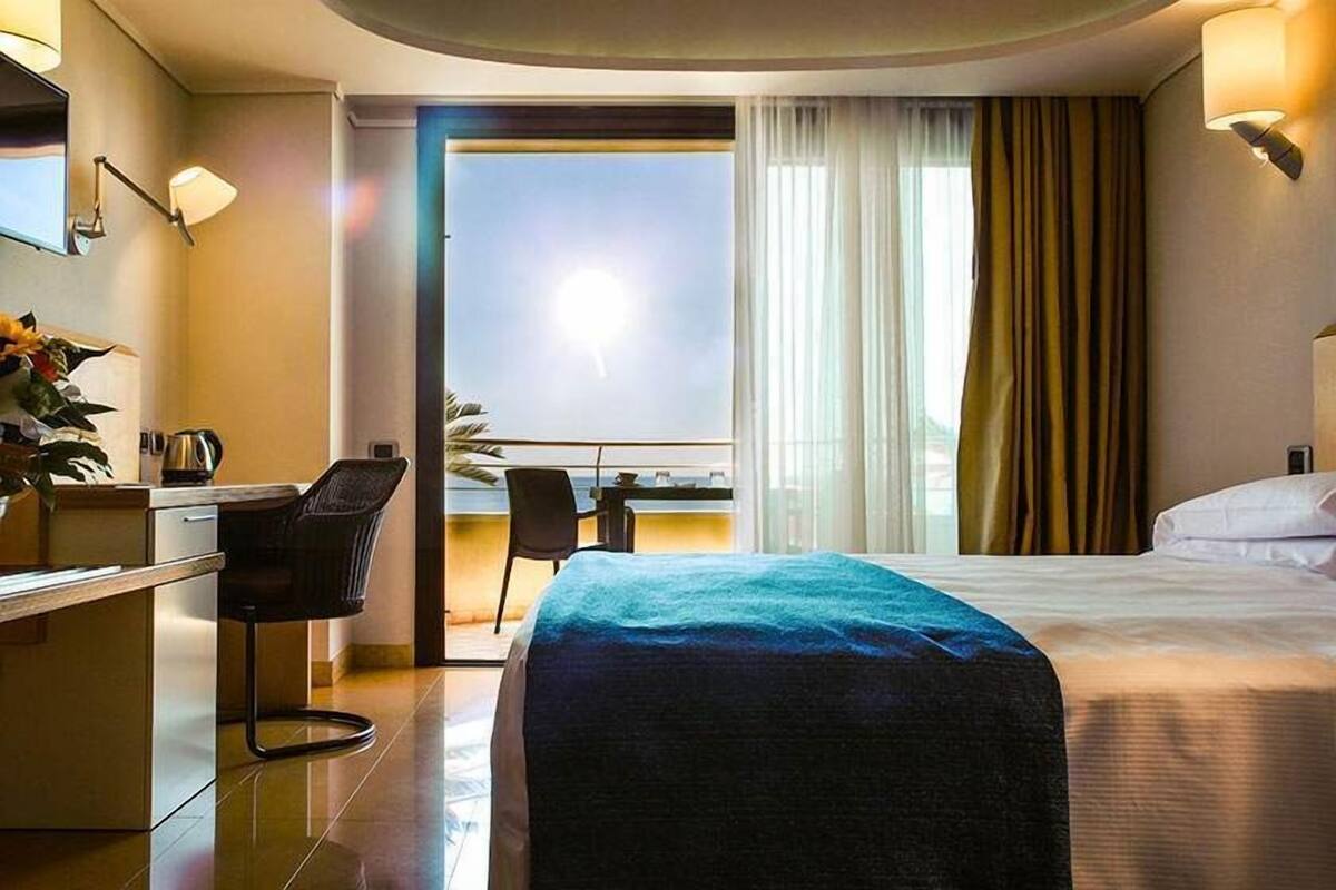 A hotel room with a bed, a desk, a chair, a small table, and a view of the outside.