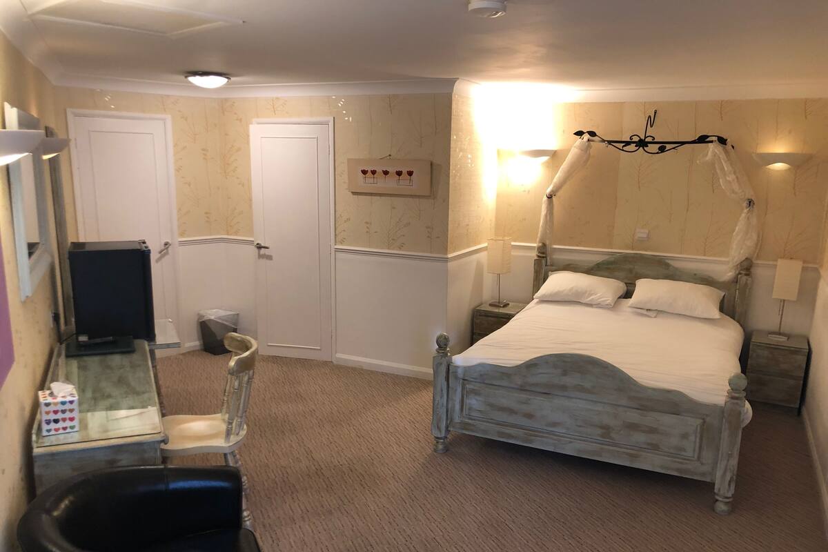 A bedroom with a four-poster bed, a bedside table, a chair, a desk with a computer, and a trash bin.