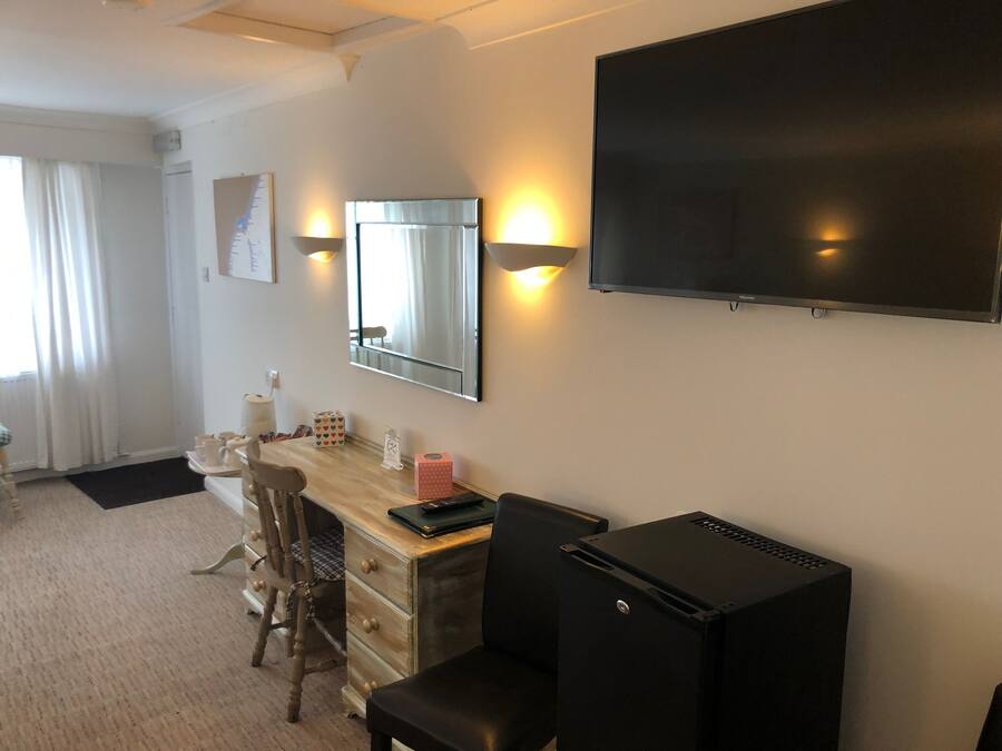A hotel room with a wooden desk, a chair, a small black refrigerator, a flat-screen TV mounted on the wall, and a window with curtains.