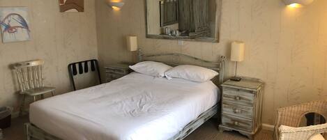 A neatly made bed with white linens, a wooden nightstand with a lamp, a wicker chair, and a mirror above the bed.
