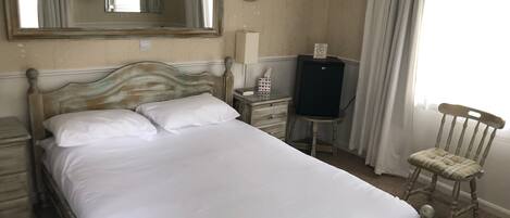 A neatly made bed with white linens, a wooden headboard, a bedside table with a lamp, a small table with a television and a tissue box, and a chair with a cushion.