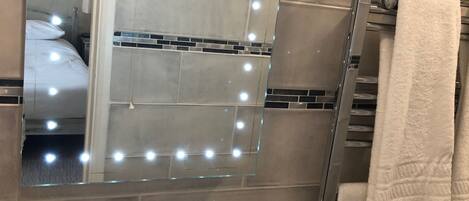 A bathroom with a vanity, a mirror with lights, a towel rack, and a shower.