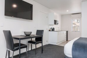 A modern hotel room with a flat-screen TV, a dining area with a table and chairs, a kitchenette, and a bed with a blue accent. - Braemar Motor Lodge (Palmerston North)