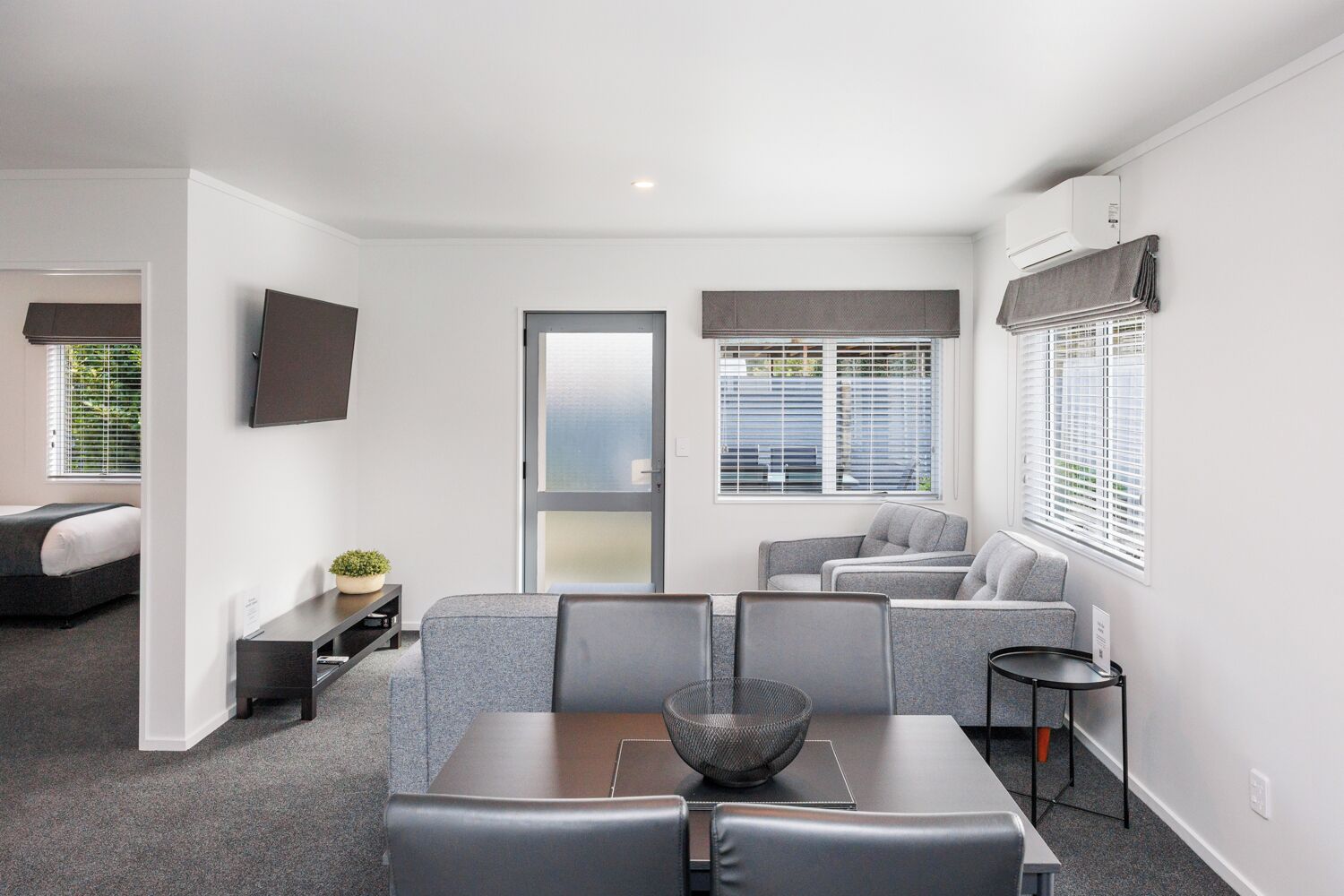 Et moderne hotelværelse med spiseplads, en sofa, et fjernsyn på væggen og et stort vindue med skodder.