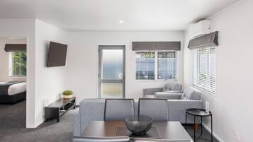 Et moderne hotelværelse med spiseplads, en sofa, et fjernsyn på væggen og et stort vindue med skodder.