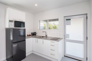 A compact kitchen with a refrigerator, microwave, sink, and a window with blinds. - Braemar Motor Lodge (Palmerston North)