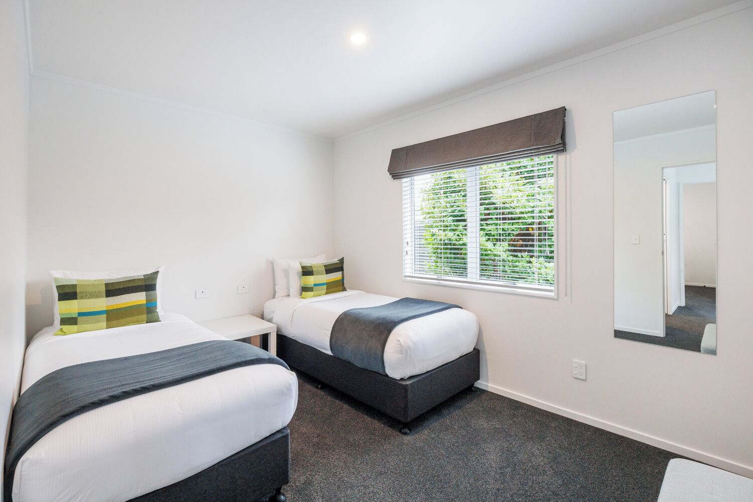 A twin room with two beds, a window with blinds, a mirror, and a carpeted floor.