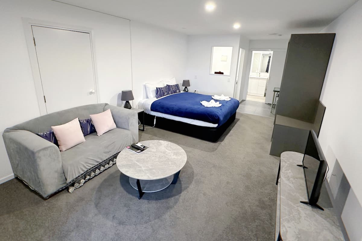 A modern hotel room with a grey sofa, a round coffee table, a large bed with a blue cover, and a flat-screen TV.