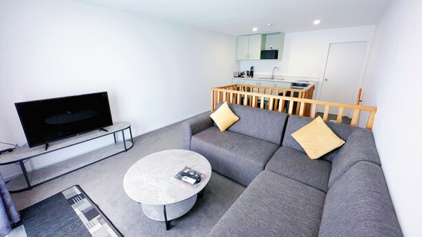 A modern living room with a grey sofa, a coffee table, a flat-screen TV, and a kitchen area in the background. - Brylin Motel (Rotorua)