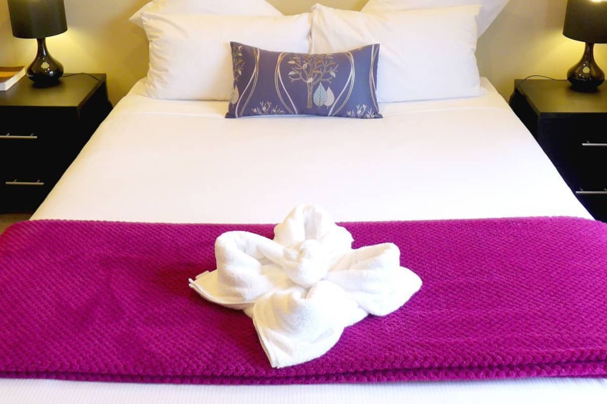 A hotel room with a bed, two bedside lamps, a framed picture on the wall, and a purple throw blanket.