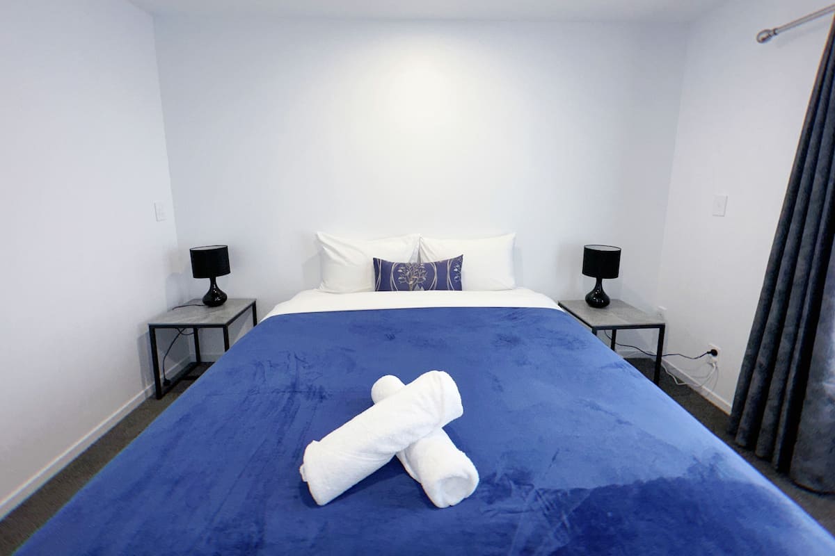 A hotel room with a large bed, two bedside tables with lamps, a blue bedspread, and a white towel on the bed.