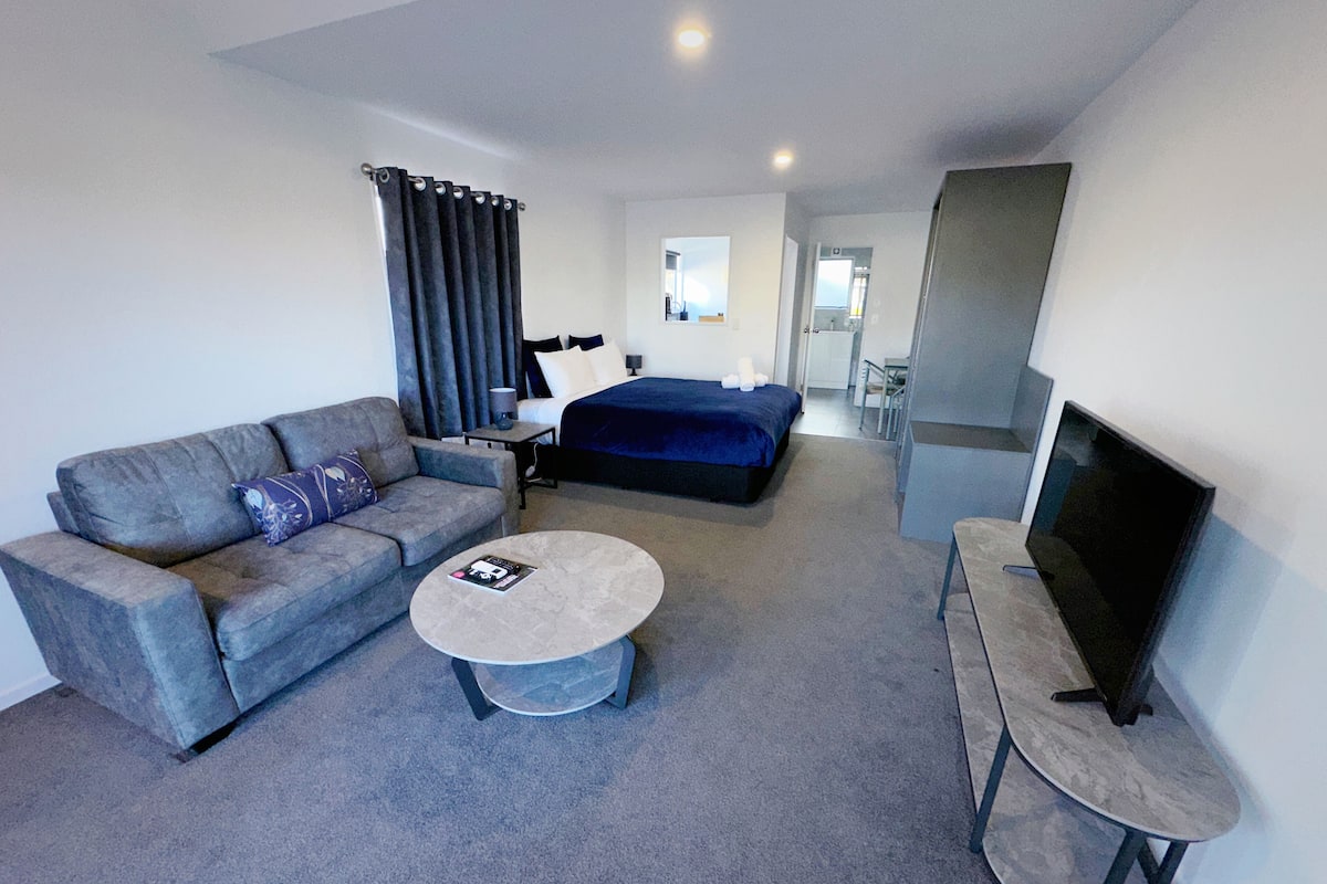 A modern hotel room with a grey sofa, a round coffee table, a bed with a blue duvet, a TV on a stand, and a bathroom visible in the background.