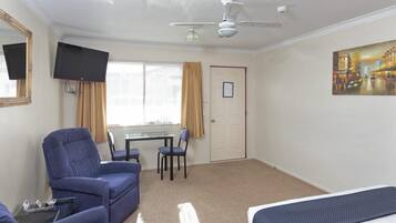 Habitación de hotel con cama, escritorio con silla, sillón azul, televisor y ventilador de techo.