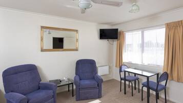 A room with two blue armchairs, a glass-top table with four chairs, a ceiling fan, a flat-screen TV, a large mirror, and a window with curtains.