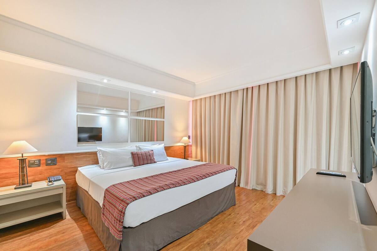 Una habitación de hotel con cama, lámparas de noche, televisor, espejo y ventana con cortinas.
