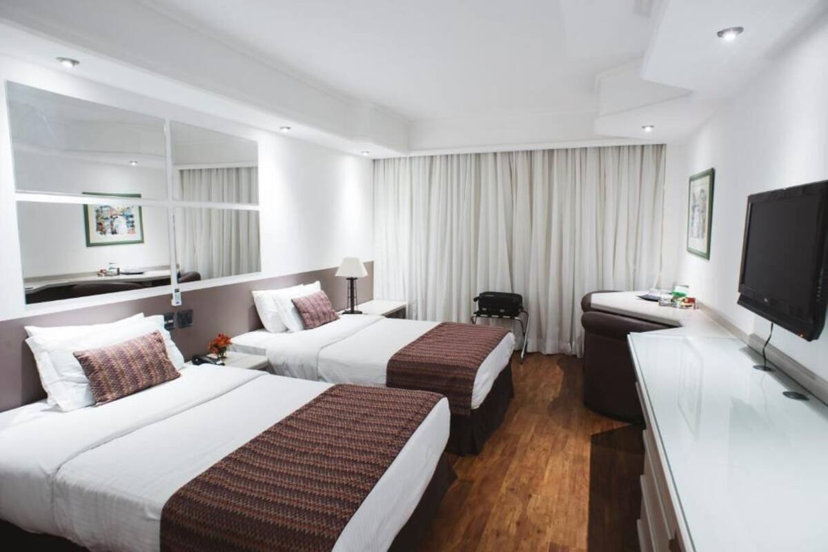 Habitación de hotel con dos camas, un escritorio, una silla, un televisor y un ventanal con cortinas.