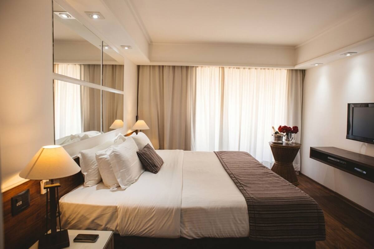 Habitación de hotel con cama, lámparas de noche, televisión, espejo y ventana con cortinas.