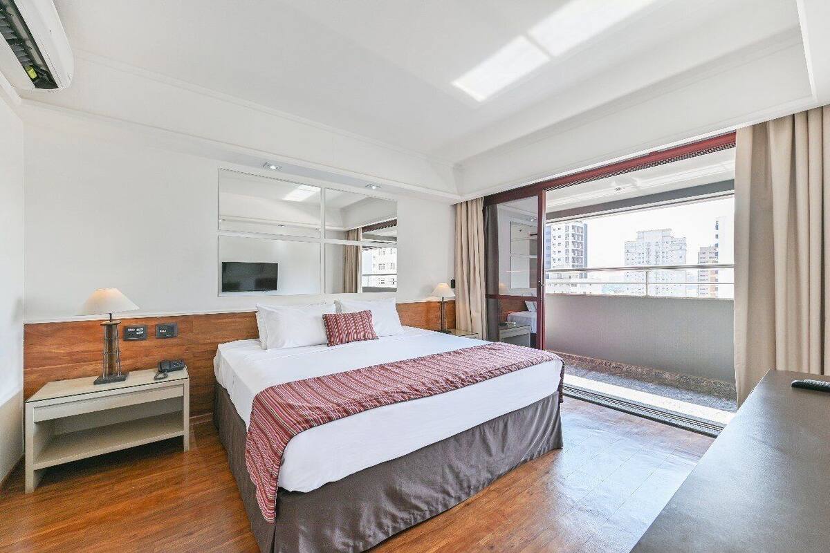 Habitación de hotel con cama, mesitas de noche, televisor, balcón con vista a la ciudad y ventana con cortinas.