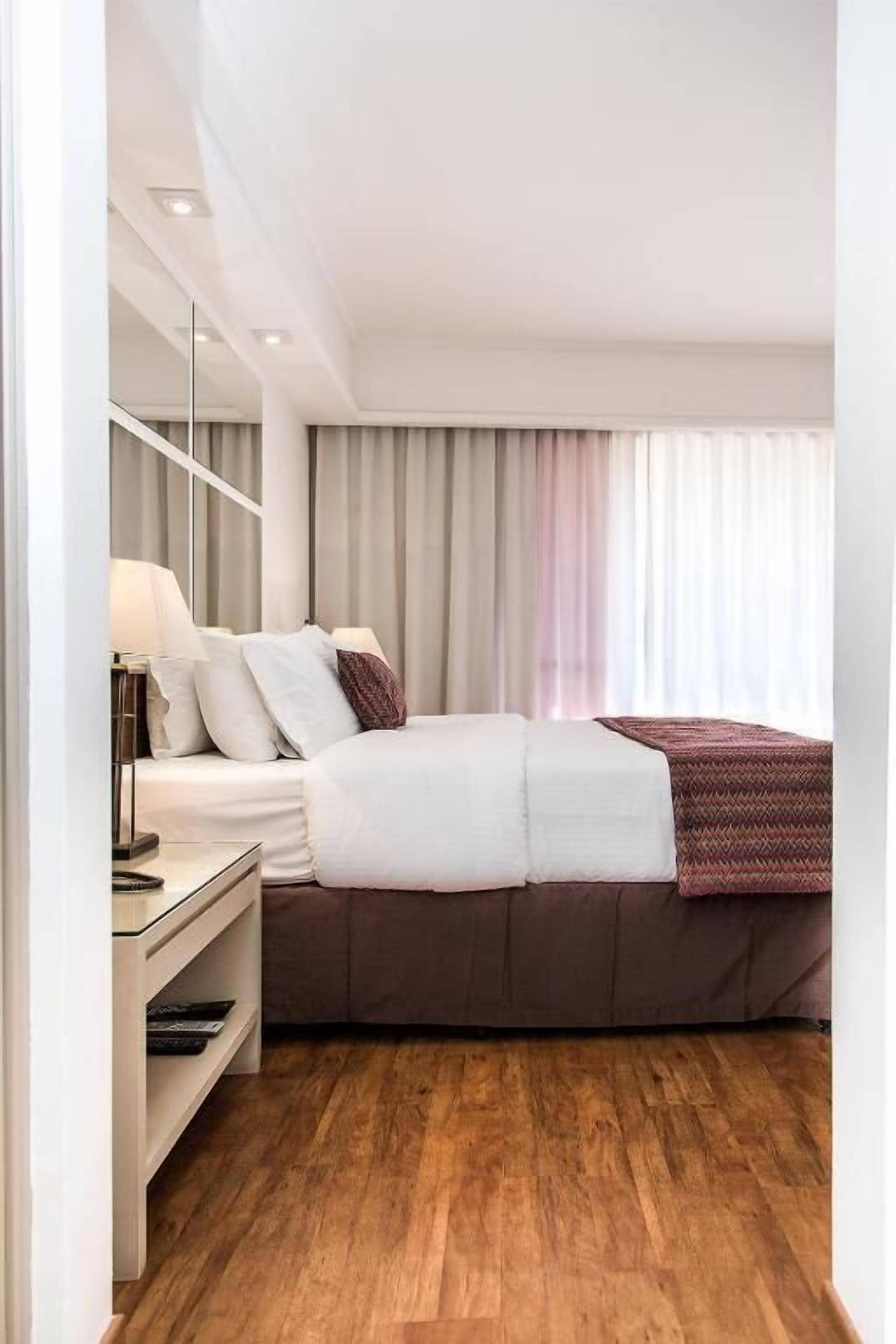 A modern hotel room with a bed, bedside table, lamp, and a view of the outside through curtains.