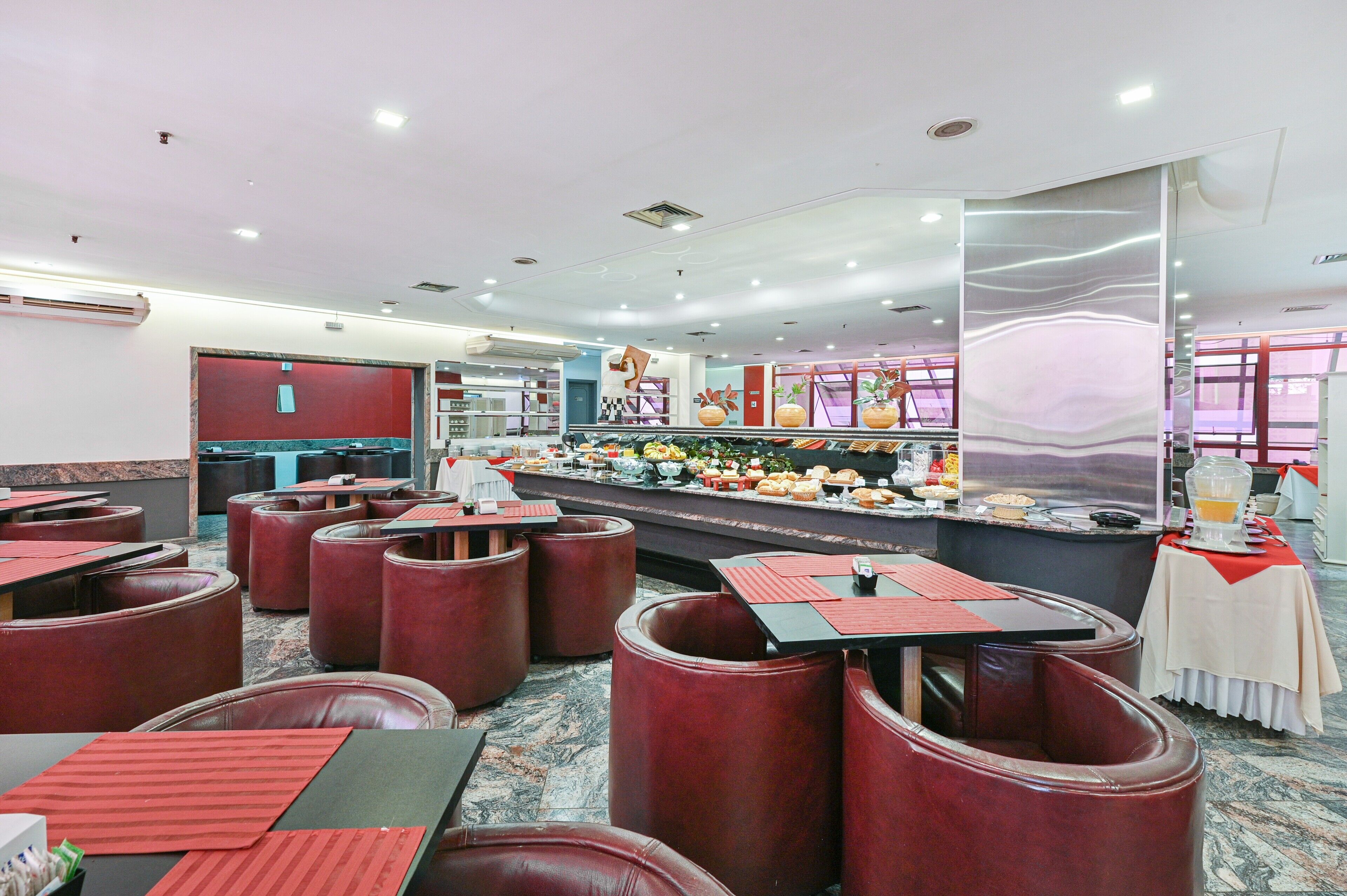 Um restaurante com cabines e mesas, um buffet com diversos pratos e um balcão com dispensador de bebidas.