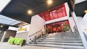 Entrada de hotel con escaleras, un área techada y una ventana iluminada en rojo.