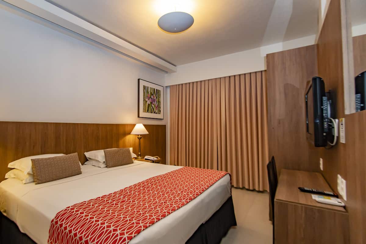 Ein Hotelzimmer mit einem großen Bett, zwei Nachttischlampen, einem hölzernen Kopfteil, einem an der Wand befestigten Fernseher und einem Fenster mit Vorhängen.
