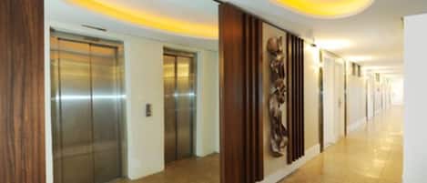 A modern hotel corridor with elevators, a wooden panel wall, and a polished floor.