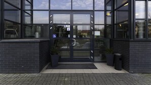 Une entrée moderne avec une porte en verre, un panneau indiquant l’accessibilité pour les personnes à mobilité réduite et deux plantes en pot.