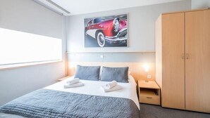 Une chambre d’hôtel avec un grand lit, deux oreillers, une table de chevet, une lampe, une armoire et un tableau représentant une voiture, accroché au mur.