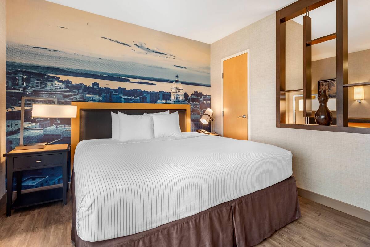 Habitación de hotel con una cama grande, una mesita de noche con lámpara, un mural de paisaje urbano en la pared y una ventana con vistas a la ciudad.