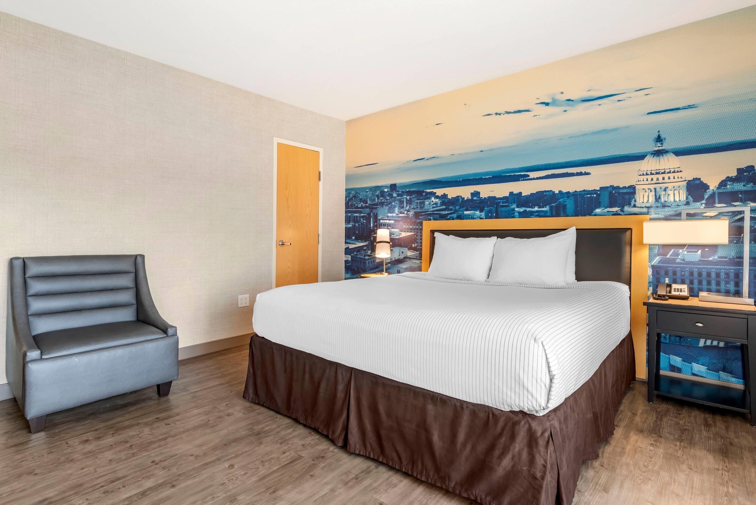 Ein Hotelzimmer mit einem großen Bett, einem grauen Sessel, einem Nachttisch mit Lampe und einem Stadtbild-Wandgemälde.