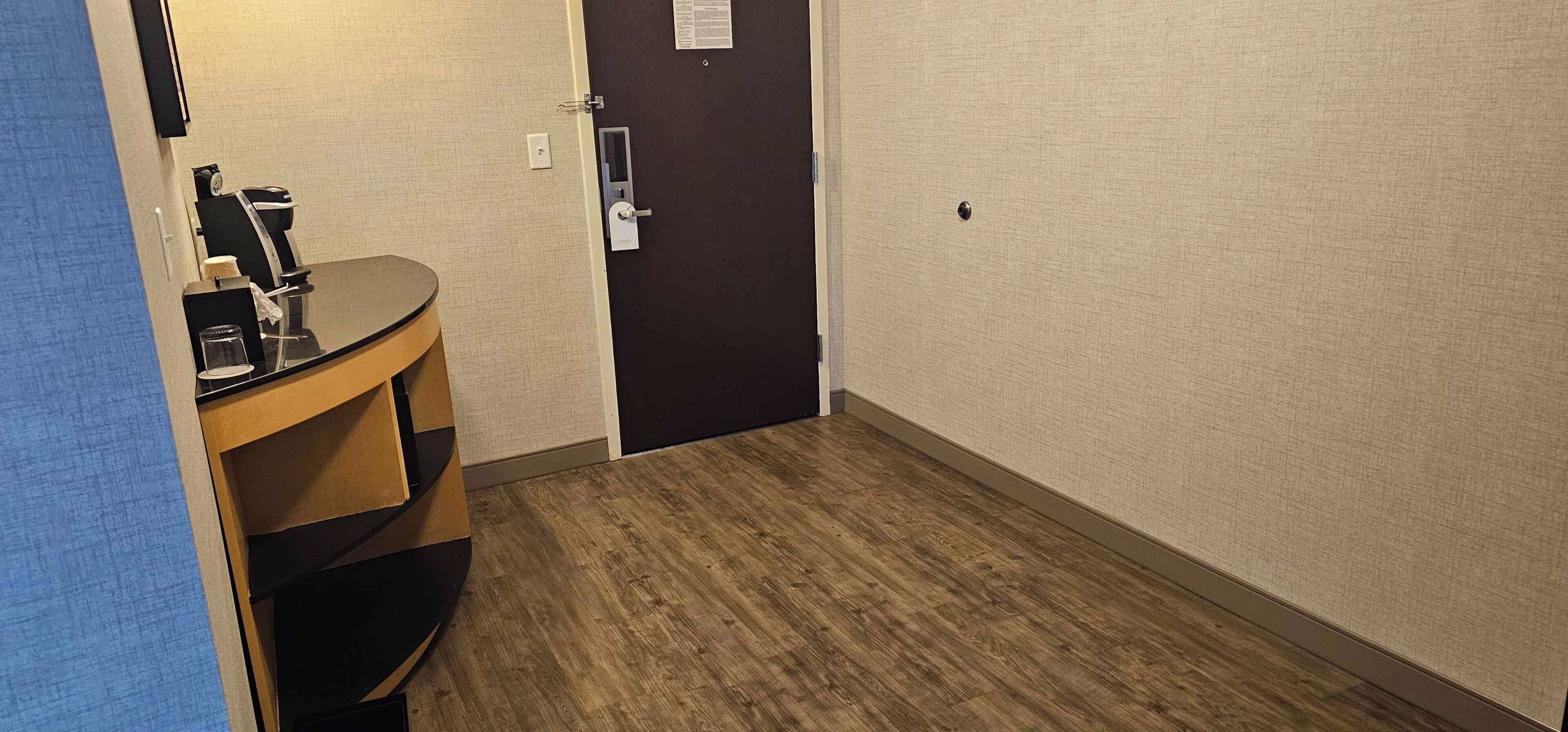 A hotel room with a wooden floor, a black door, a small round table with a coffee maker, and a mirror.