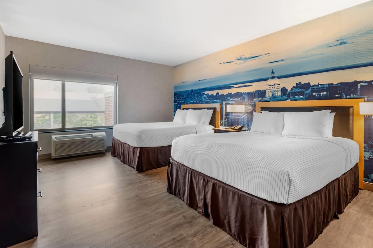 Habitación de hotel con dos camas, un ventanal grande, un mural de la ciudad y un escritorio con una computadora.