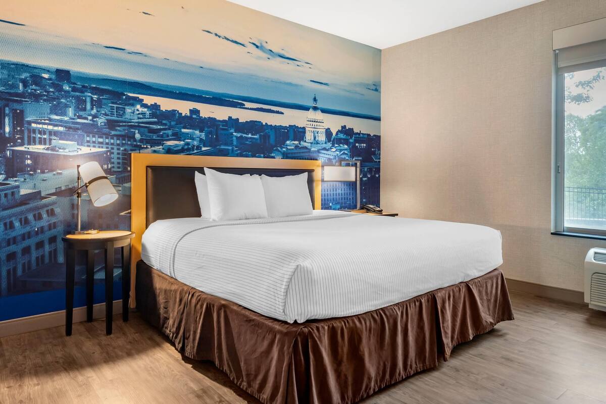 Habitación de hotel con una cama grande, una mesita de noche, una lámpara y un mural con vista a la ciudad.